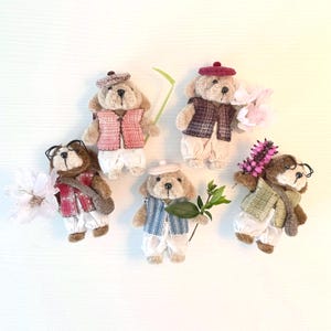 Puede incluir: Cinco osos de peluche en miniatura vestidos con diferentes atuendos, incluyendo chalecos y boinas. Los osos sostienen flores o ramitas. Están dispuestos sobre una superficie blanca, creando una escena encantadora y caprichosa.