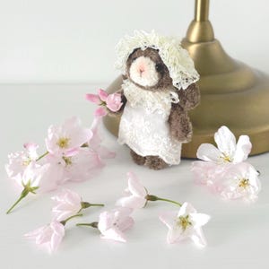 May include: A small, brown teddy bear wearing a white lace dress and bonnet, holding pink flowers. Delicate pink cherry blossoms are scattered around the bear. The bear is standing near a gold-colored lamp base.