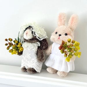 May include: Two plush bunny figurines, one brown and one cream-colored, dressed in white dresses and holding yellow floral bouquets. The brown bunny wears a lace bonnet. The bunnies are displayed on a white shelf against a white background.