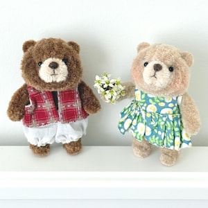 Puede incluir: Dos osos de peluche. El oso de la izquierda es marrón oscuro, viste un chaleco de cuadros rojos y pantalones blancos. El oso de la derecha es de color canela claro, lleva un vestido azul con estampado floral blanco y amarillo y sostiene flores.