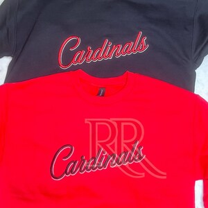 May include: A black crewneck sweatshirt and a red crewneck sweatshirt, both featuring the word Cardinals embroidered in a cursive script font with white and red outlines across the center of the chest area on each garment.