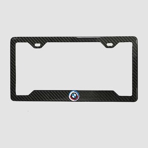 May include: A black carbon fiber license plate frame with the BMW logo in the lower right corner. The frame has a rectangular shape with rounded corners and mounting holes at the top.