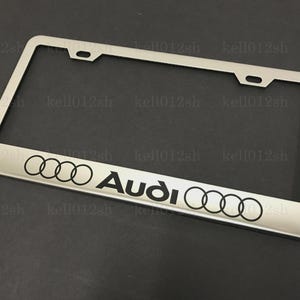 May include: Silver-colored license plate frame with the Audi logo in black. The frame has a rectangular shape with rounded corners and mounting holes. The Audi logo consists of four interlocking rings and the brand name.