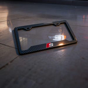 May include: Carbon fiber license plate frame with a glossy finish. The frame has a black and gray woven pattern and features a red and white "S" logo. The frame is designed to fit a standard U.S. license plate.