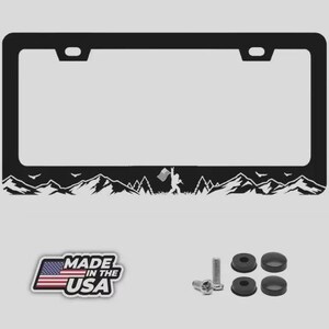 May include: Black license plate frame with a mountain and Bigfoot silhouette design. Includes mounting hardware and a "Made in the USA" sticker. The frame has a rectangular shape with rounded corners.