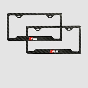 May include: Two carbon fiber license plate frames. Each frame has a black carbon fiber weave pattern and a red and white "RS" logo. The frames are rectangular with rounded corners and mounting holes.