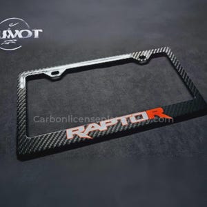 May include: A carbon fiber license plate frame with the word "RAPTOR" in white and orange lettering. The frame has a black and gray checkered pattern and is rectangular with mounting holes.
