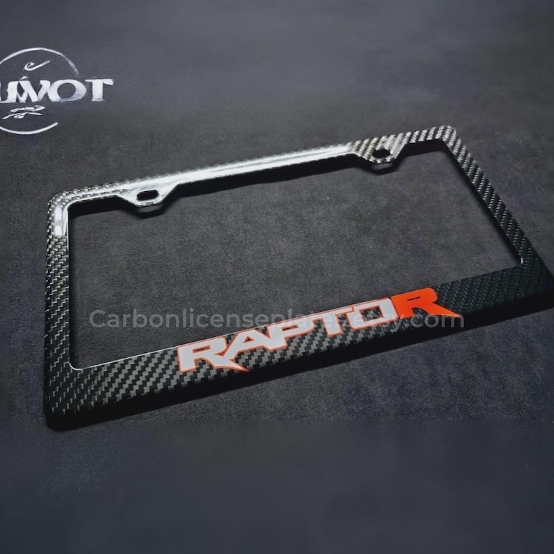 May include: A carbon fiber license plate frame with the word "RAPTOR" in white and orange lettering. The frame has a black and gray checkered pattern and is rectangular with mounting holes.