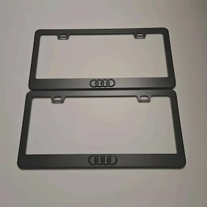 Puede incluir: Dos marcos de matrícula negros mate. Cada marco tiene el logotipo de Audi en el centro inferior. Los marcos tienen orificios de montaje en la parte superior y son rectangulares.