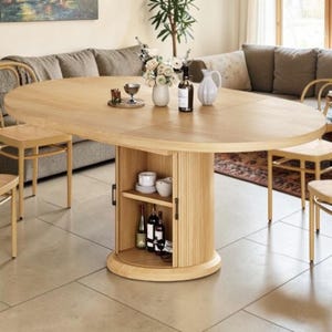 May include: Oval wooden dining table with a built-in storage cabinet. The table is light-colored, with a natural wood grain finish. The cabinet has shelves for storing items like wine bottles and dishes. The table is surrounded by wooden chairs.