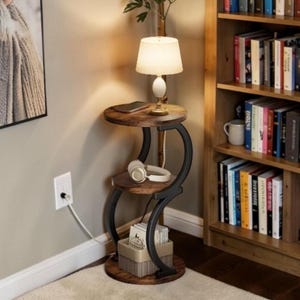 May include: A three-tiered side table with a lamp, headphones, and a small basket. The table has a dark brown wooden top and base with black metal supports. A small lamp with a cream-colored shade sits on the top tier.
