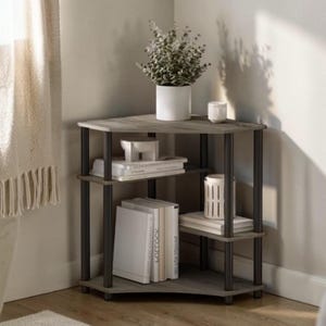 May include: A gray corner shelf unit with three tiers, holding books, decorative items, and a potted plant. The shelves have a wood-grain appearance, supported by dark metal legs. A white curtain is visible in the background.