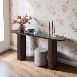 May include: A dark wood console table with a dark gray oval top, fluted legs, and a small gray upholstered stool. The table is decorated with a vase of dried leaves, a small dish, and gold candle holders with purple candles. The background features floral wallpaper.