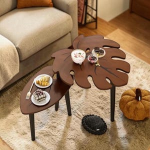 May include: A unique, brown leaf-shaped coffee table with black legs. The table has two tiers and is decorated with small plates of food and a cup of coffee. A decorative pumpkin and a small black pillow are on the floor.