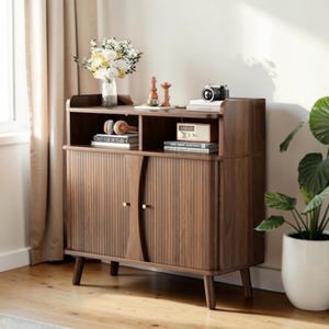 May include: A dark wood cabinet with a tambour door design, featuring an open top shelf and two lower doors with small gold knobs. The cabinet stands on four tapered legs and holds a vase of flowers, a camera, and several books on its surface.