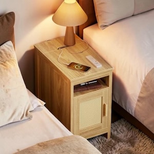 May include: A light wood bedside table with a lamp, phone, and remote. The table has a door with a woven design and a shelf. A phone is charging on the top. The lamp has a beige lampshade.
