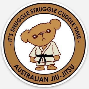 May include: A circular sticker with a brown border and a white interior. It depicts a cartoon teddy bear in a martial arts uniform with a black belt. Text around the bear reads: "IT'S SNUGGLE STRUGGLE CUDDLE TIME - AUSTRALIAN JIU-JITSU."
