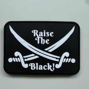 Puede incluir: Parche rectangular negro con texto blanco y espadas cruzadas. El texto dice "Raise The Black!" en una fuente estilizada. Las espadas son blancas y curvas, con empuñaduras detalladas.