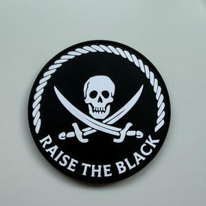 May include: A circular black patch with a white skull and crossed swords emblem. The words "RAISE THE BLACK" are written in white around the edge, enclosed by a white rope-style border.