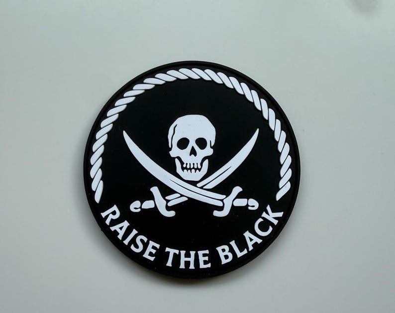 Op de afbeelding: Een ronde, zwarte patch met een wit doodshoofd en gekruiste zwaarden. De tekst "RAISE THE BLACK" staat in het wit rond de rand, omlijst door een witte touwrand.