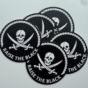 Könnte beinhalten: Eine Sammlung schwarzer und weißer runder Aufkleber. Jeder Aufkleber zeigt einen Totenkopf mit Piraten-Augenklappe und gekreuzten Schwertern, umgeben von einem weißen Seilrand. Der Text "RAISE THE BLACK" ist in Weiß gedruckt.