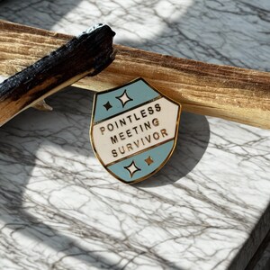 May include: A gold-rimmed enamel pin with a shield design. The pin features a light blue and white background with the text "POINTLESS MEETING SURVIVOR". Two diamond shapes are on the top and bottom of the pin.