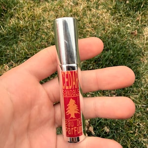 May include: A silver perfume bottle with a red label displaying the words "CEDAR SANDALS" and a tree graphic. The bottle is labelled "eau de parfum". The bottle is held in an open hand against a backdrop of green grass.
