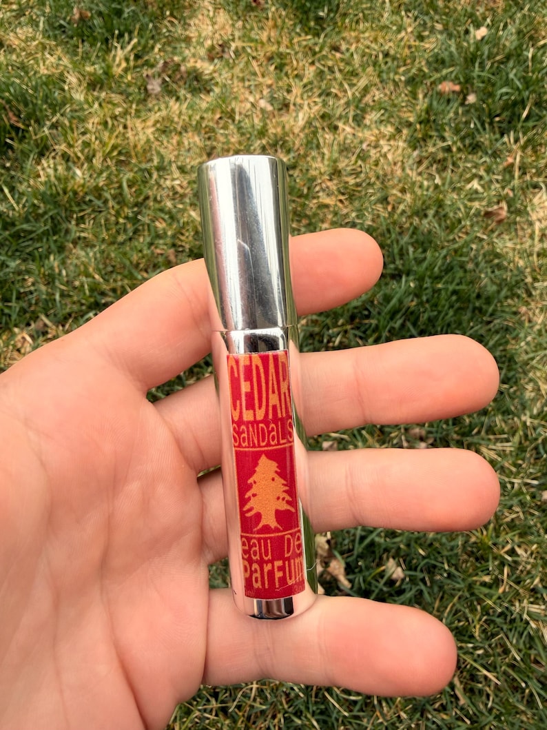 May include: A silver perfume bottle with a red label displaying the words "CEDAR SANDALS" and a tree graphic. The bottle is labelled "eau de parfum". The bottle is held in an open hand against a backdrop of green grass.