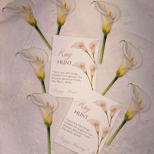 May include: Two rectangular white cards with the text Ring HUNT, There Are 100 Rings Hidden Throughout The Shower, Whoever Finds The Most, WINS! and Happy Hunting! surrounded by six white and yellow calla lily flower illustrations.