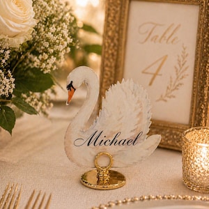 Könnte beinhalten: Eine weiße, schwanenförmige Platzkarte mit dem Namen Michael in schwarzer Schreibschrift. Der Schwan hat einen orangefarbenen Schnabel und ist auf einem kleinen, runden, polierten goldfarbenen Metallständer auf einem Tisch platziert.