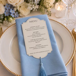 May include: A cream-colored menu card with a decorative scalloped edge and black text, featuring a light blue tassel attached at the bottom. The menu is placed on a folded light blue cloth napkin, which rests on a white plate with a gold beaded rim.