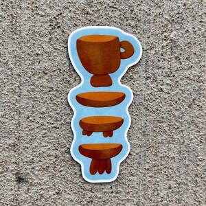 May include: A sticker featuring a brown coffee cup above three brown tables. The sticker has a light blue outline and is on a textured gray surface. The design is a whimsical illustration.