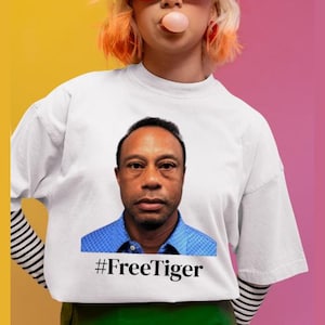 Puede incluir: Camiseta blanca con un retrato de un hombre con camisa azul y el texto "#FreeTiger" debajo. La camiseta la lleva una persona con pelo naranja y rubio, y gafas de sol rojas.