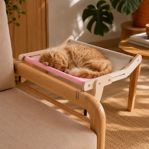 Könnte beinhalten: Ein Katzenbett aus Holz mit einer rosa-cremefarbenen Hängematte. Eine flauschige, orangefarbene Katze schläft zusammengerollt in der Hängematte. Das Bett ist an einem beige gepolsterten Stuhl befestigt.