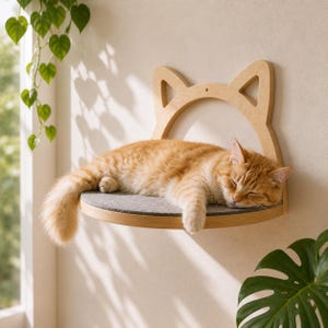 Könnte beinhalten: Ein hölzernes Katzenregal in Form von Katzenohren, mit einer schlafenden, orangefarbenen Tabbykatze. Das Regal hat ein graues Filzpolster. Die Katze liegt zusammengerollt auf dem Regal, die Augen geschlossen. Das Regal ist an einer beigen Wand befestigt.
