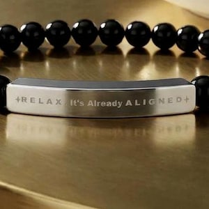 May include: Black beaded bracelet with a silver bar inscribed with the words "+RELAX It's Already ALIGNED+". The bracelet is on a gold-colored surface, with a white and gold dish in the background.