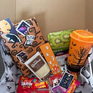 May include: A cardboard gift box filled with assorted items including an orange reusable cup with a black tree silhouette, a packet of Carnation hot chocolate, Biscoff biscuits, Skittles, Starburst, tea, bookmarks, and stickers featuring Halloween themes.
