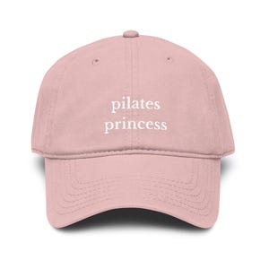 Peut inclure: Casquette de baseball rose clair avec l'inscription "pilates princess" en blanc. La casquette a une visière incurvée et une couronne arrondie. Un accessoire élégant pour les amateurs de fitness.