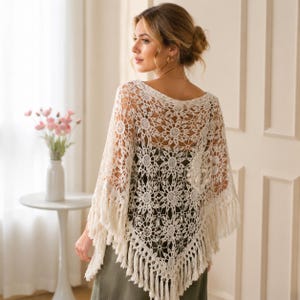 May include: A cream-colored crochet poncho featuring an intricate floral pattern throughout the fabric. The garment has a wide neckline and is finished with long, dangling fringe along the bottom edges, creating a textured and layered appearance.