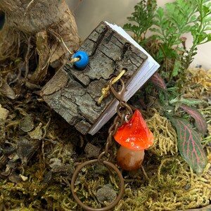 May include: A miniature book with a textured, dark brown bark cover, bound with yellow string and a single blue bead. Attached to the binding is a brown metal keychain featuring a glossy, bright orange mushroom charm with a tan stem.