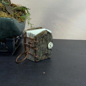 May include: A miniature book keychain featuring a cover made of textured brown tree bark. The pages are white and stacked. The binding is secured with thin, multi-colored braided cord and a bronze-toned metal ring. A small white button is attached.