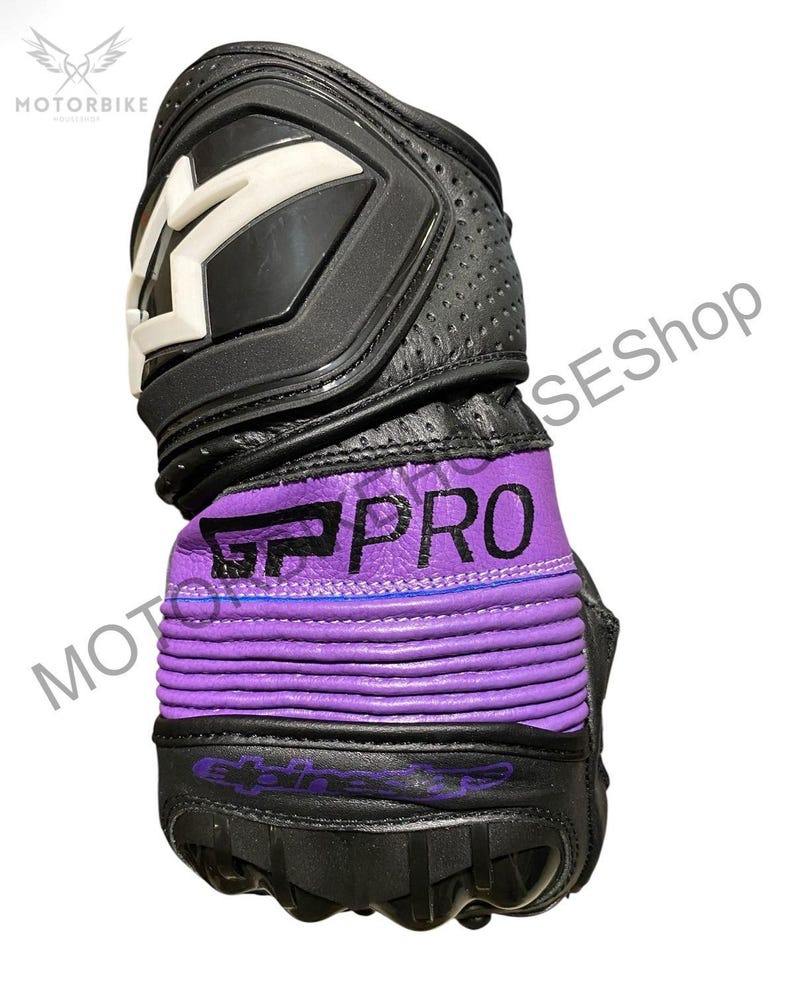 Puede incluir: Guante de moto negro y morado con detalles blancos. El guante presenta el texto "GP PRO" en blanco sobre una banda morada. La parte superior tiene un panel protector negro con un dise&ntilde;o de "M" blanco. Se ve cuero perforado.