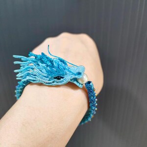 May include: A blue dragon bracelet with intricate woven details. The dragon's head features a pearl and black accents. The bracelet is a vibrant shade of blue, with a detailed design. The bracelet is a unique piece of jewelry.