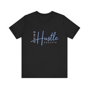 Può includere: T-shirt nera con la parola "Hustle" in corsivo azzurro chiaro. Le parole "Grind" ed "Execute" sono sopra e sotto "Hustle". La maglietta ha un girocollo ed è realizzata in materiale morbido.