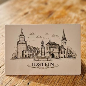 May include: An off-white rectangular card with a black and white illustration of Idstein, Germany. The drawing depicts a tower, buildings, a fountain, and a church. The word "IDSTEIN" is written below the illustration.