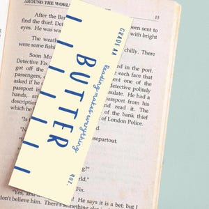 May include: A cream-colored bookmark with the word "BUTTER" in large blue letters. The text "Reading makes everything" is written vertically. The bookmark is placed in an open book with text visible.
