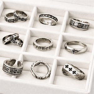 May include: A collection of silver rings with various designs, including cross motifs, scrollwork, and engraved text. The rings are displayed in a white jewelry box, showcasing their intricate details and craftsmanship. Some rings feature the text "FOREVER" and "ONE HEART".