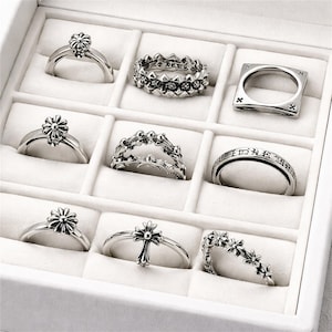 May include: A collection of silver rings displayed in a white jewelry box. The rings feature various designs, including floral, cross, and geometric patterns. Some rings have engraved text, and all are set against a white background.