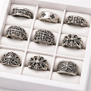Op de afbeelding: Een collectie van negen zilveren ringen met diverse sierlijke ontwerpen, waaronder kruisen, bloempatronen en geometrische vormen. De ringen worden gepresenteerd in een witte doos met individuele compartimenten, die de ingewikkelde details tonen.