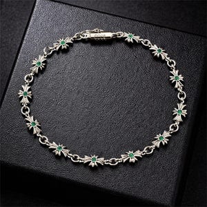Puede incluir: Pulsera de plata con eslabones en forma de cruz, cada uno adornado con una pequeña gema verde redonda en el centro. La pulsera se muestra sobre una superficie negra. El cierre dice "1983".